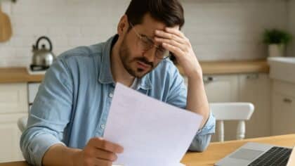 Un homme fatigué et préoccupé quant à ses indémnités lisant des documents financiers à sa table de cuisine, sans texte sur l’image.